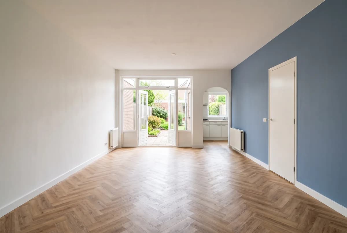Lege woonkamer van een jaren-70/80-woning: brede pui naar de tuin, doorgang naar de dichte keuken